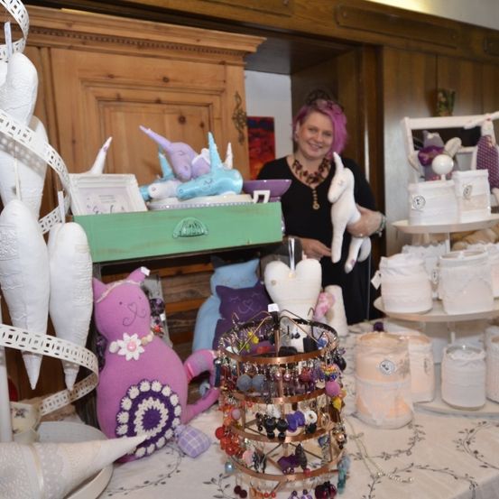 Eine Frau mit rosa Haaren steht hinter einem Tisch voller verschiedener Gegenstände. Auf dem Tisch befinden sich Gegenstände wie Kuscheltiere, Kerzen und ein dekorativer Korb. Im Hintergrund steht auch ein Regal mit weiteren Kuscheltieren und Dekorationsartikeln.