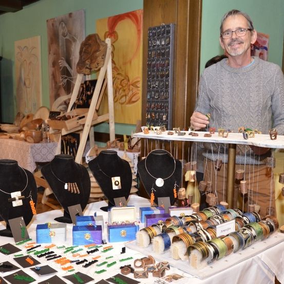 Ein Mann steht an einem Tisch, auf dem verschiedene Schmuckstücke wie Halsketten und Armbänder ausgestellt sind. Hinter ihm sind verschiedene Kunstwerke an der Wand aufgehängt, und zusätzliche Gegenstände sind auf dem Tisch angeordnet.