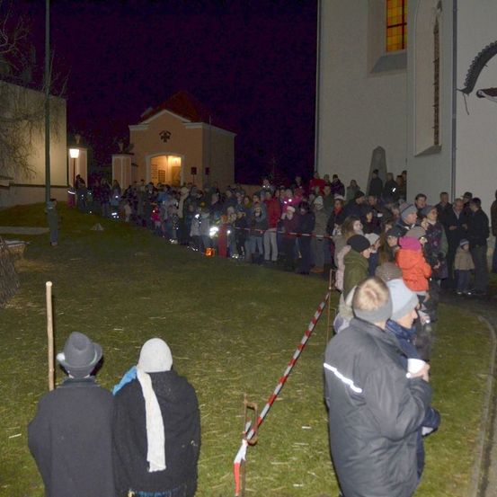 Bei Nacht versammelt sich eine Menschenmenge vor einer Kirche, trägt Winterkleidung und Mützen. Eine rot-weiße Barriere trennt sie vom Gebäude. Einige halten Taschenlampen.