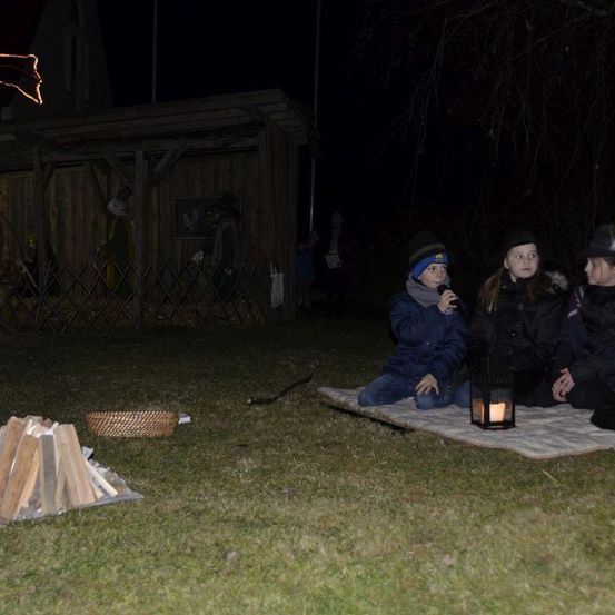 Drei Kinder sitzen auf einer Decke in der Nähe einer brennenden Laterne und einem Haufen Holz. Dahinter steht ein kleines Holzhaus, und Menschen stehen davor.