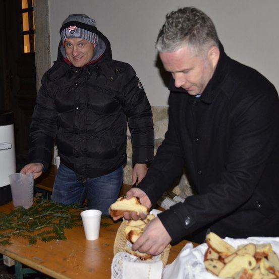Zwei Männer stehen an einem Tisch, einer mit einem Stück Brot, der andere greift nach mehr. Hinter ihnen ist eine weiße Wand und eine Tür.