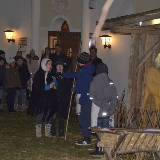 Eine Gruppe von Menschen versammelt sich im Freien in der Nähe einer Krippe, einige halten Kerzen und andere lauschen einem Sprecher mit Mikrofon.