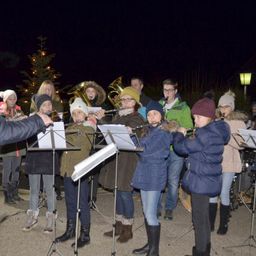Eine Gruppe von Menschen, möglicherweise eine Schulband, spielt nachts auf einer Bühne Instrumente. Sie tragen Winterkleidung und einige haben Mützen. Hinter ihnen steht ein Weihnachtsbaum.