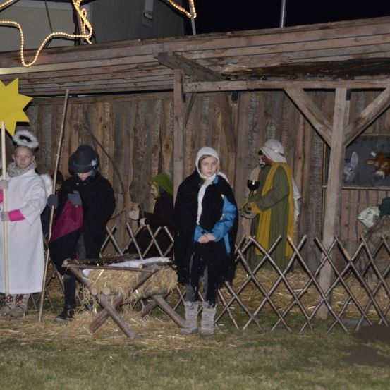 Eine Krippenszene wird nachts im Freien aufgeführt. Mehrere in Kostümen gekleidete Personen stehen um eine Krippe mit Heu herum. Ein Stern hängt über ihnen, und Tiere sind im Hintergrund sichtbar.