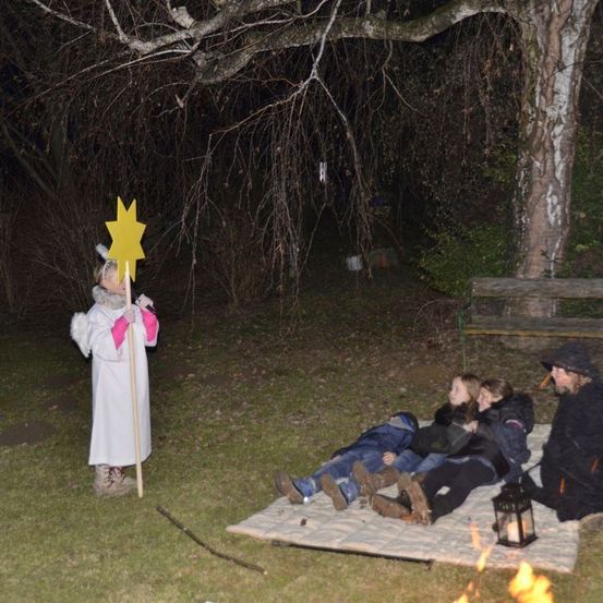 Eine Gruppe von Menschen sitzt auf einer Decke in der Nähe eines Feuers. Eine Person in einem weißen Gewand mit einem Stern an einem Stock steht neben ihnen. Das Gebiet ist dunkel.