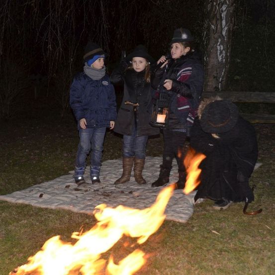 Vier Kinder versammeln sich in der Nacht um ein Lagerfeuer. Eines hält eine Laterne und ein anderes ein Mikrofon, während ein weiteres Kind in die Hocke geht.