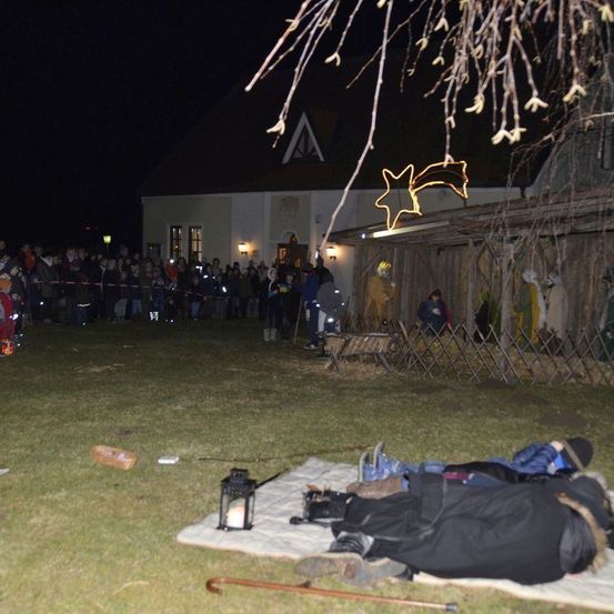 Eine Menschenmenge versammelt sich bei Nacht für eine Krippenszene vor einer Kirche, mit Lichtern und Dekorationen.