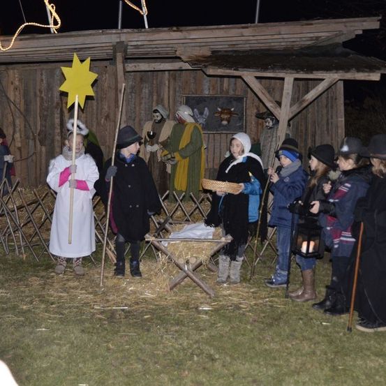 Eine Gruppe von Kindern in Kostümen steht um eine Krippe herum, eine mit einem Stern und andere mit Laternen. Die Szene befindet sich vor einer Holzstruktur.