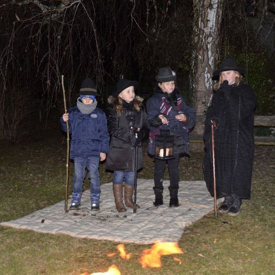 Vier Personen stehen in der Nähe eines Lagerfeuers, möglicherweise in einem Wald. Drei tragen Hüte und Stiefel. Eine hält eine Laterne, und eine andere hält einen Stock.