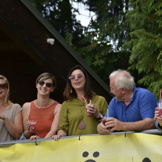 Eine Gruppe Erwachsener, die lächelnd für ein Foto posieren, Getränke in der Hand halten und hinter einem gelben Banner mit schwarzen Pfotenabdrücken stehen. Bäume sind hinter ihnen.