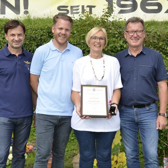Vier Erwachsene stehen lächelnd zusammen in einem Garten. Eine Frau in der Mitte hält ein gerahmtes Diplom. Hinter ihnen steht ein Banner mit der Aufschrift Seit 1967.