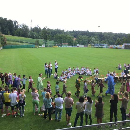 Eine Gruppe von Menschen versammelt sich auf einem Feld mit Bäumen im Hintergrund. Einige Leute stehen, andere sitzen auf dem Gras. Einige Leute tragen Hüte und Schuhe. Eine Person trägt einen blauen Umhang. Ein Mann spielt eine Trommel. Es gibt mehrere Werbebanner am Zaun.