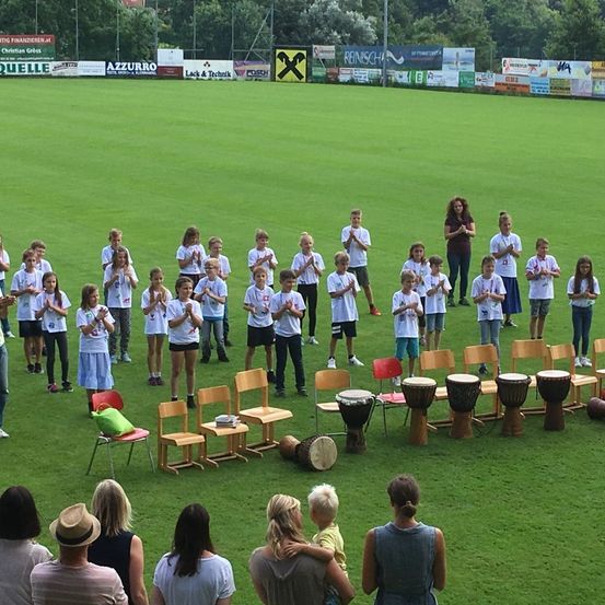 Eine Gruppe von Kindern in weißen T-Shirts und Shorts ist auf einem Rasenfeld versammelt und führt eine synchronisierte Klapproutine durch. Trommeln und Stühle sind um sie herum angeordnet. Vor ihnen beobachten Erwachsene. Im Hintergrund sind Banner angebracht.