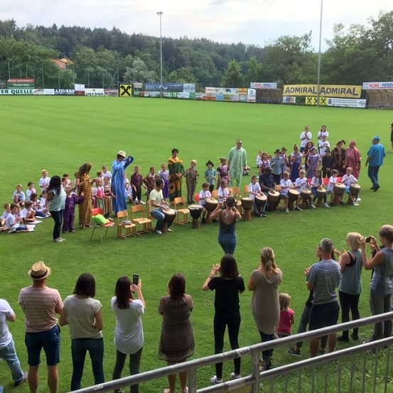 Eine Menschenmenge versammelt sich auf einem Feld für eine Aufführung, wobei Kinder in einem Kreis sitzen und Erwachsene um sie herum stehen. Die Kinder tragen Kostüme und spielen Trommeln. Werbung ist im Hintergrund sichtbar.