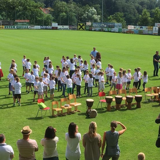Eine Gruppe von Kindern in weißen Hemden und Shorts steht auf einem grünen Feld, hält sich an den Händen und steht im Kreis. Es gibt Trommeln auf dem Boden und Stühle um sie herum. Einige Erwachsene beobachten sie.