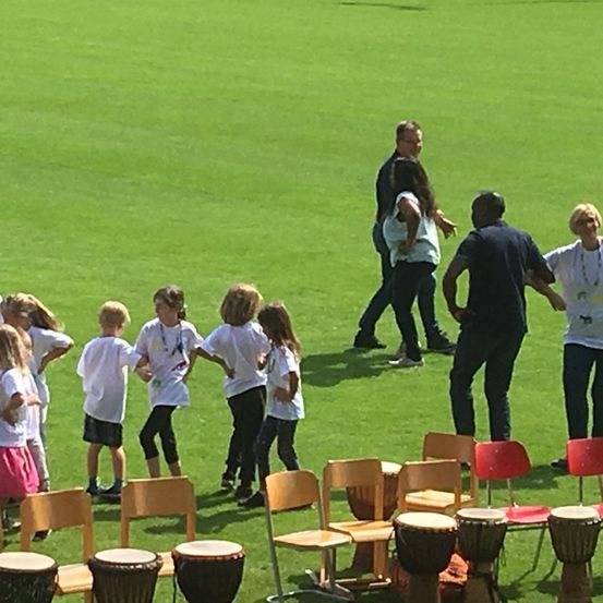 Eine Gruppe von Kindern und Erwachsenen befindet sich auf einem grünen Feld und praktiziert möglicherweise einen Tanz. Sie halten sich an den Händen, während die Erwachsenen hinter ihnen stehen. Vor ihnen stehen Trommeln und Stühle.