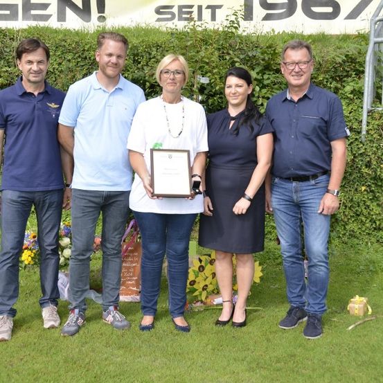Fünf Personen stehen auf einem Rasenfeld und lächeln für ein Foto. Die Frau in der Mitte hält einen gerahmten Preis. Hinter ihnen sind Blumen und eine Banner mit Text zu sehen.