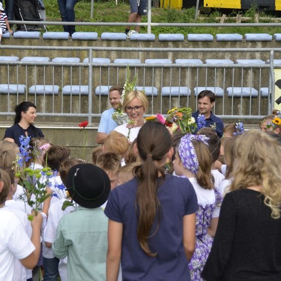 Eine Menschenmenge mit Blumen in einem Stadion, darunter eine Frau mit Brille und eine Gruppe von Kindern.