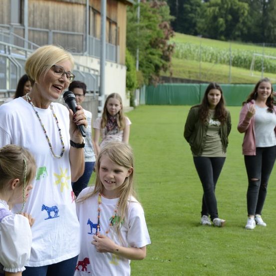 Eine ältere Frau spricht in ein Mikrofon, während eine Gruppe junger Mädchen zuhört. Sie befinden sich auf einem Feld mit einem Stadion im Hintergrund.