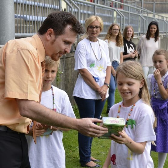 Ein Mann in einem orangefarbenen Hemd übergibt einem Mädchen eine Schachtel. Andere Kinder und Frauen schauen zu. Sie stehen auf Gras mit einer Wand und Tribünen dahinter.