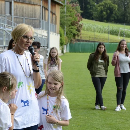 Eine ältere Frau mit einem Mikrofon spricht zu zwei jungen Mädchen mit bemalten Pferden auf ihren Shirts. Sie stehen auf einem Feld mit einem Gebäude und Bäumen im Hintergrund.