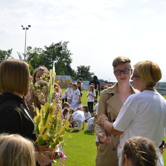 Eine Gruppe von Menschen versammelt sich auf einem Rasenfeld. Ein Mann mit Brille und Trenchcoat steht zwischen zwei Frauen. Eine Frau hält einen Blumenstrauß. Im Hintergrund hockt und steht eine Gruppe von Kindern, und ein Zaun mit Bannern ist sichtbar.