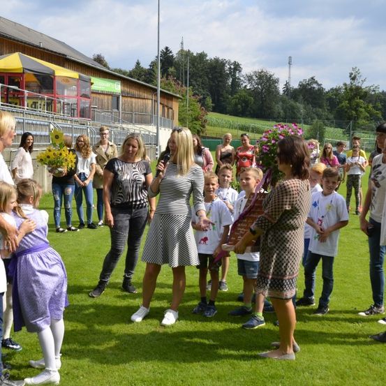 Eine Gruppe von Menschen, darunter Kinder, steht auf einem Feld mit einem Gebäude im Hintergrund. Eine Frau hält ein Mikrofon und scheint zu sprechen. Eine andere Frau hält einen Strauß Sonnenblumen. Einige Leute halten Blumen. Der Himmel ist klar.