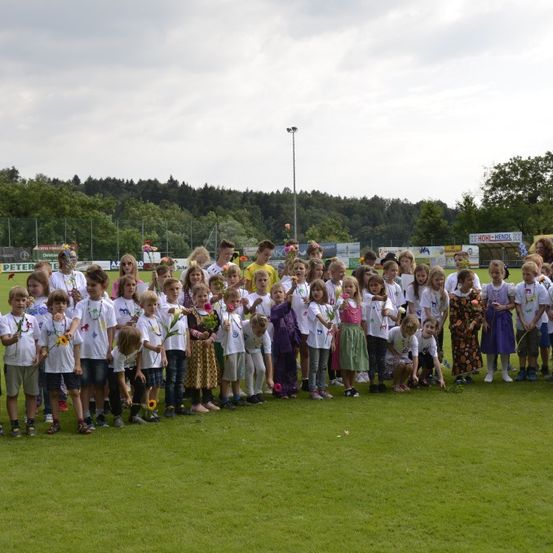 Eine große Gruppe von Kindern in weißen Hemden und mit Blumen posiert für ein Foto auf einem Rasenfeld mit Bäumen und einem bewölkten Himmel im Hintergrund.