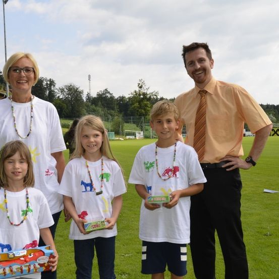 Eine Gruppe von vier Kindern und zwei Erwachsenen posiert auf einem Rasenfeld. Sie tragen alle weiße Hemden und halten eine Schachtel mit Spielzeug. Die Erwachsenen lächeln.