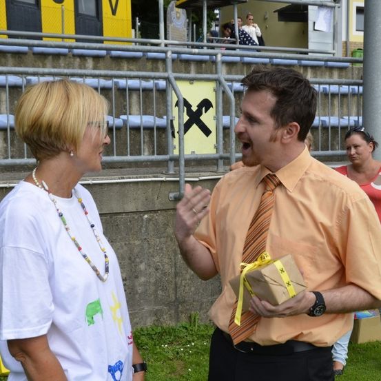 Ein Mann in einem orangefarbenen Hemd, der ein Geschenk in der Hand hält, spricht mit einer Frau in einem weißen T-Shirt. Sie stehen in einem Außenbereich in der Nähe eines Zauns. Im Hintergrund sind ein Gebäude und Personen zu sehen.