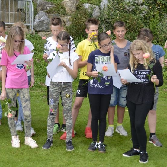 Eine Gruppe junger Menschen versammelt sich auf einer Wiese, hält Papiere und Blumen in den Händen und nimmt wahrscheinlich an einer Schulveranstaltung teil. Sie sind lässig gekleidet.