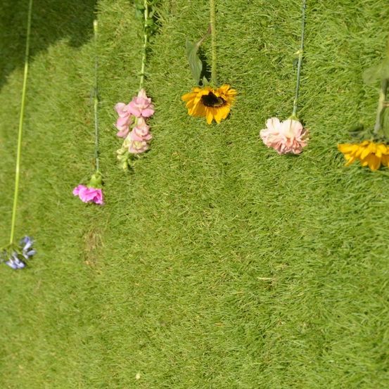 Eine Gartenszene mit verschiedenen Blumen, die an grünen Stielen auf einem Grasfeld hängen. Es gibt rosa, gelbe und blaue Blumen mit Blättern.