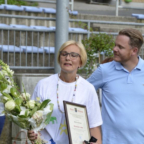 Eine Frau in einem weißen T-Shirt hält einen Blumenstrauß und ein gerahmtes Zertifikat, während sie neben einem Mann in einem blauen Hemd steht. Sie sind von einer Außenumgebung mit einem Stadion und grünen Pflanzen im Hintergrund umgeben.