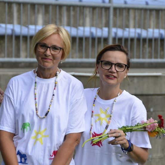 Zwei Frauen stehen nebeneinander vor einem leeren Stadion, tragen weiße T-Shirts mit bunten Tieraufdrucken und halten einen Blumenstrauß.
