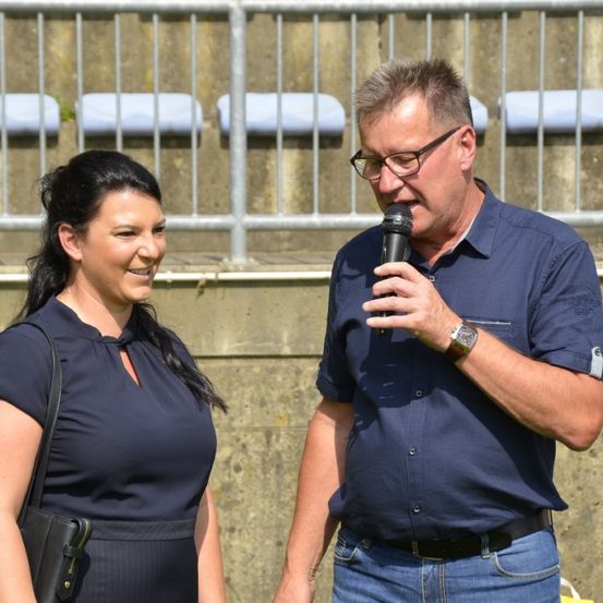 Ein Mann und eine Frau stehen lächelnd vor einem Zaun. Der Mann hält ein Mikrofon und scheint zu sprechen.