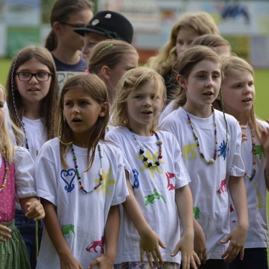 Eine Gruppe junger Mädchen in weißen T-Shirts mit bunten Drucken steht in einer Reihe, einige tragen Halsketten, möglicherweise bei einer Schulveranstaltung.
