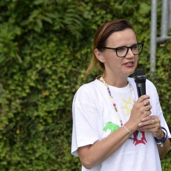 Eine Frau mit Brille spricht in ein Mikrofon. Sie trägt ein weißes T-Shirt mit buntem Design. Hinter ihr ist ein verschwommener Hintergrund mit grünem Laub.