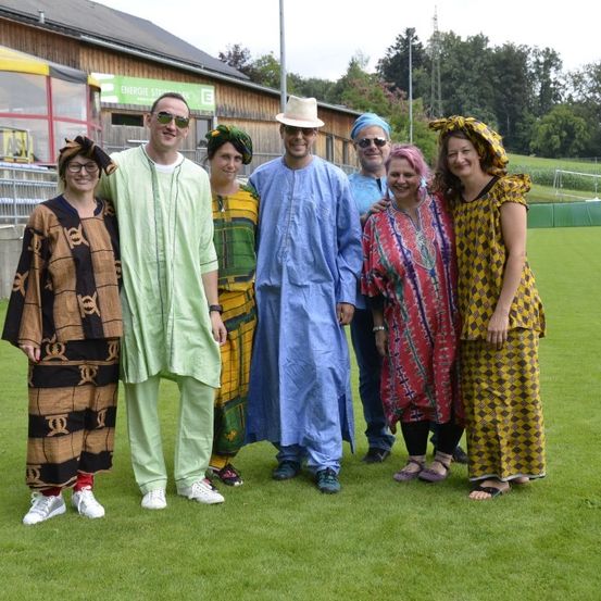 Sieben Personen stehen auf einem grünen Rasen, gekleidet in farbenfrohe afrikanische Kleidung, mit Gebäuden und Grünpflanzen im Hintergrund.