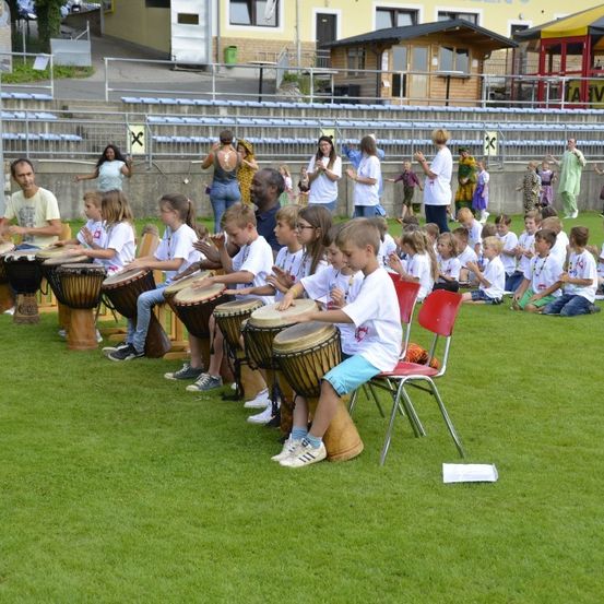 Kinder in weißen Shirts und Shorts spielen Trommeln im Freien, in der Nähe von Erwachsenen und Tribünen im Hintergrund.