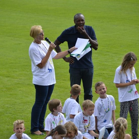 Eine Gruppe von Kindern und zwei Erwachsenen befindet sich auf einem Rasenfeld. Eine Frau mit Brille hält ein Mikrofon und Papier. Der Mann hält ein Stück Papier und berührt seinen Mund. Die Kinder schauen sich etwas an.
