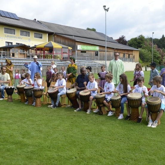 Eine Gruppe von Kindern und Erwachsenen sitzt um Trommeln auf einem Rasenfeld herum, wahrscheinlich an einer kulturellen Veranstaltung teilnehmend. Einige tragen traditionelle Kleidung. Im Hintergrund ist ein Gebäude mit Solarpanelen und einer Anzeigetafel zu sehen.