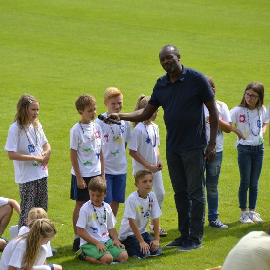 Ein Mann steht vor einer Gruppe von Kindern auf einem Feld und hält ein Mikrofon. Die Kinder tragen weiße T-Shirts mit verschiedenen Designs.