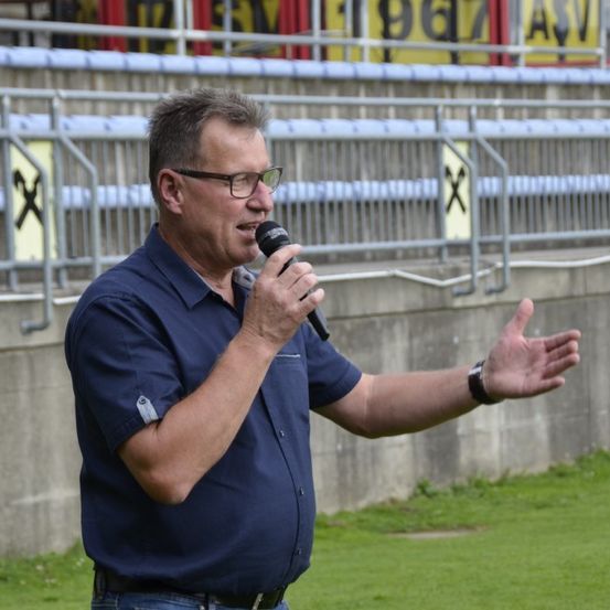 Ein Mann in einem blauen Hemd spricht in ein Mikrofon auf einem Sportfeld mit leeren Tribünen und einer Betonbarriere hinter ihm.