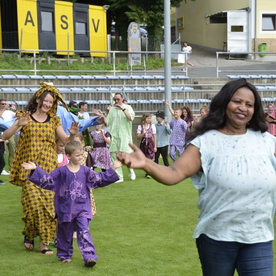 Eine Gruppe von Menschen, darunter Erwachsene und Kinder, tanzt im Freien auf einem Rasenfeld, während mehrere Menschen im Hintergrund zusehen.