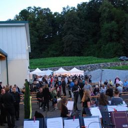 Eine Veranstaltung im Freien mit vielen Menschen, die sich um eine Bühne versammeln, umgeben von Bäumen und einer Steinmauer, mit Musikständern und einem Mikrofon aufgebaut.