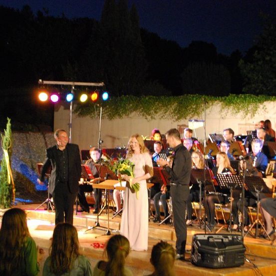 Eine Hochzeitszeremonie bei Nacht auf einer Bühne mit einer Braut, die Blumen hält, zwei Bräutigamen und einem Orchester. Das Publikum sitzt vor der Bühne.