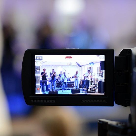 Der Kamerabildschirm zeigt eine Band, die auf der Bühne auftritt, mit Musikern, die Trompeten, Gitarren und Schlagzeug spielen. Das Wort 'AUFN' ist auf dem Bildschirm zu sehen.
