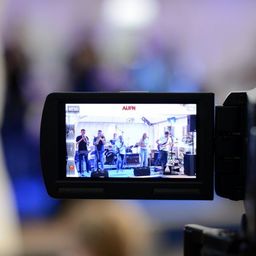 Der Kamerabildschirm zeigt eine Band, die auf der Bühne auftritt, mit Musikern, die Trompeten, Gitarren und Schlagzeug spielen. Das Wort 'AUFN' ist auf dem Bildschirm zu sehen.