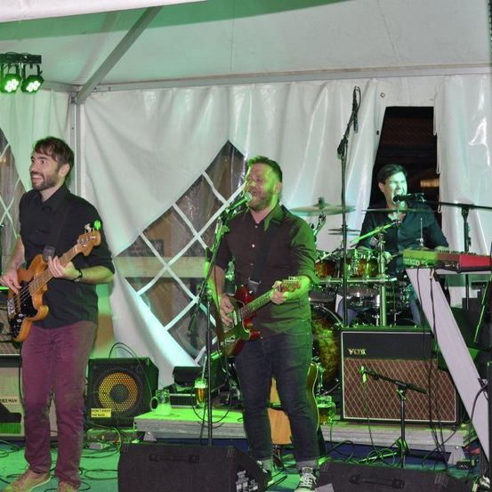 Eine Band tritt auf der Bühne unter einem Zelt auf, mit einem Mann, der Bassgitarre spielt, einem anderen, der E-Gitarre spielt, und einem Schlagzeuger hinter ihnen.