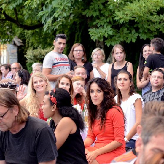 Eine Gruppe von Menschen ist im Freien versammelt, wahrscheinlich für eine Veranstaltung. Einige stehen, andere sitzen, darunter eine Frau in einem orangefarbenen Kleid. Bäume und ein Gebäude sind im Hintergrund zu sehen.